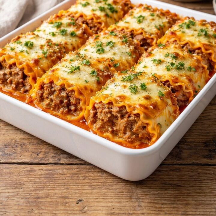 A white baking dish filled with six golden, cheese-topped short rib lasagna rolls garnished with parsley.