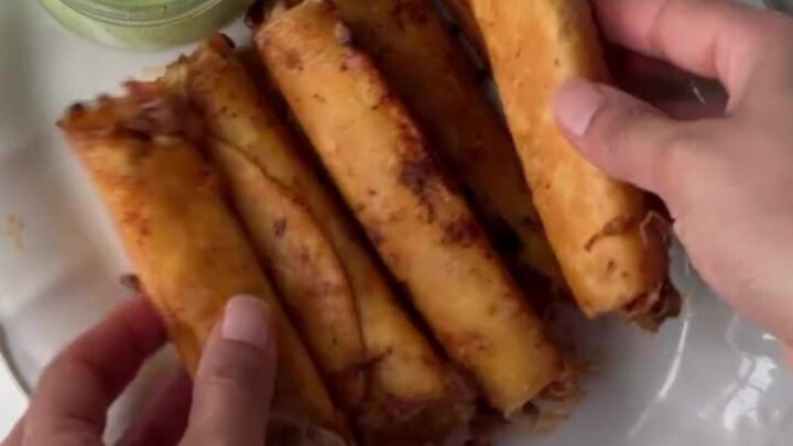 Crispy golden beef taquitos stacked on a plate with a bowl of cilantro lime crema for dipping