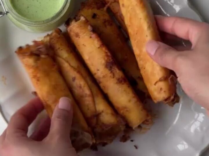 Crispy golden beef taquitos stacked on a plate with a bowl of cilantro lime crema for dipping