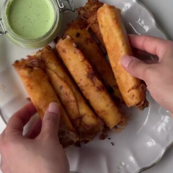 Crispy golden beef taquitos stacked on a plate with a bowl of cilantro lime crema for dipping