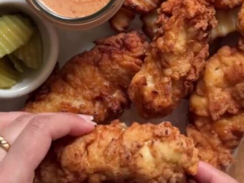 Golden crispy chicken tenders stacked on a plate with a small bowl of ranch dressing for dipping.