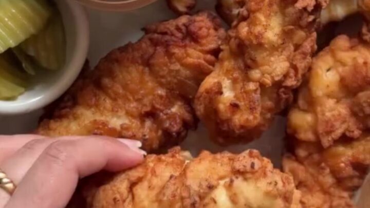 Golden crispy chicken tenders stacked on a plate with a small bowl of ranch dressing for dipping.