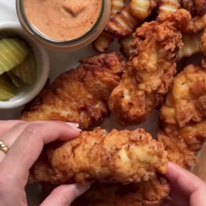 Golden crispy chicken tenders stacked on a plate with a small bowl of ranch dressing for dipping.