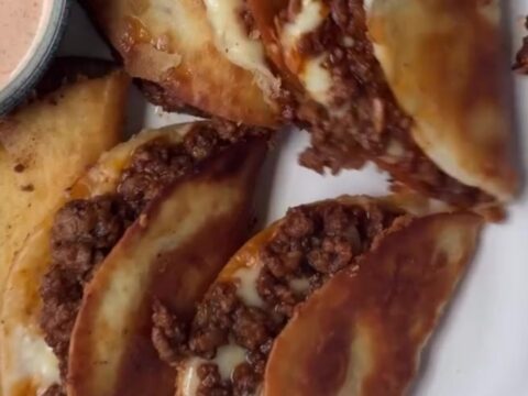 Golden crispy folded beef taco with melted cheese and pico, held by fingers, showing a crunchy bite.