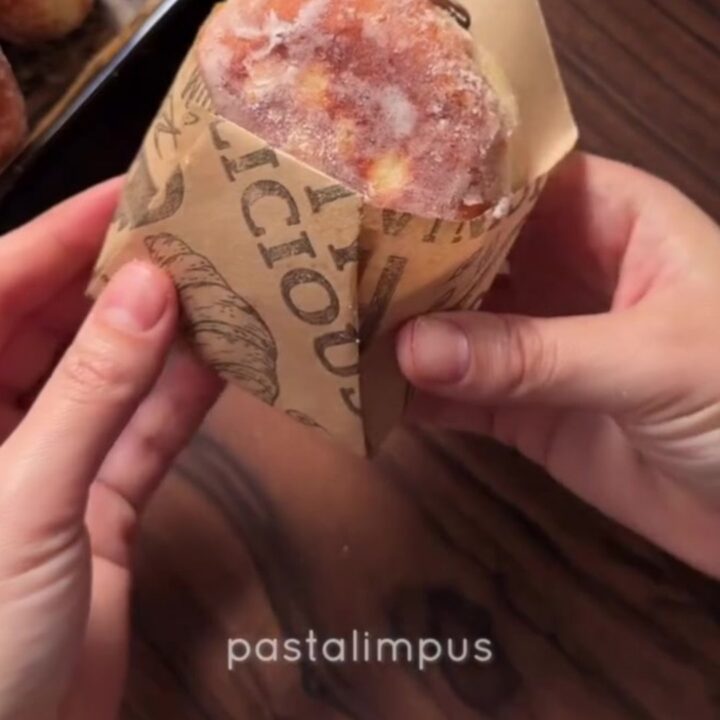 A sugar-coated donut with chocolate hazelnut filling oozing out, held in a parchment paper cone.
