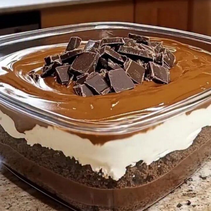 A square glass dish filled with beautiful layers of cake, whipped cream, and chocolate ganache under warm light.