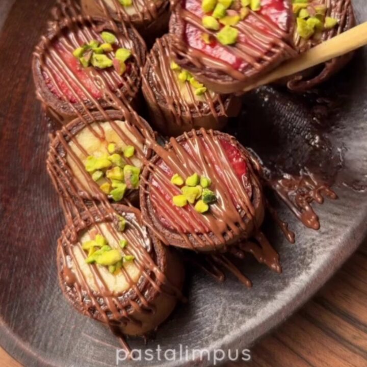Chocolate crepe sushi pieces arranged on a plate, drizzled with white chocolate and sprinkled with pistachios.