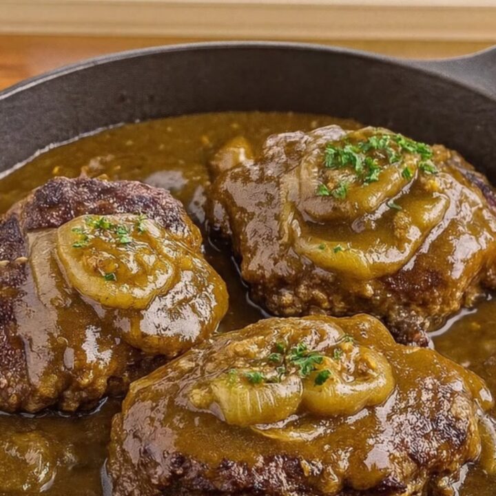 Three juicy hamburger steaks topped with caramelized onion gravy in a black cast-iron skillet.