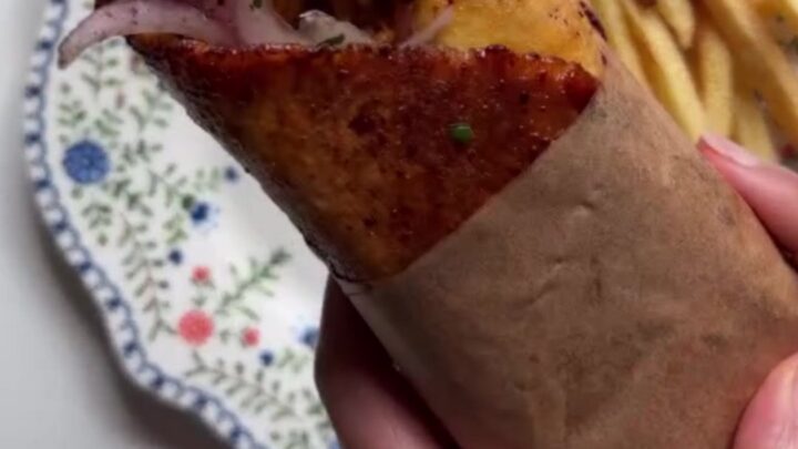 Crispy chicken shawarma wrap sliced diagonally, showing layers of chicken, fries, onions, and tomatoes, with a bowl of toum