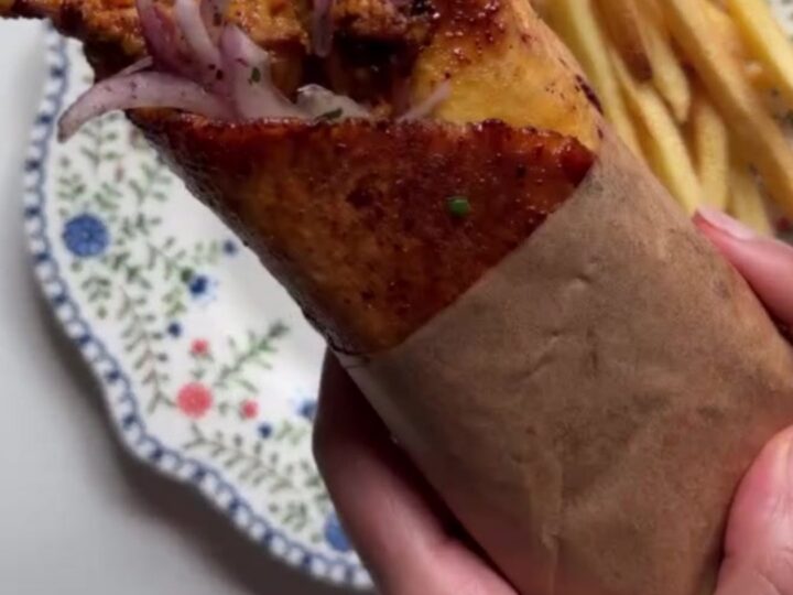 Crispy chicken shawarma wrap sliced diagonally, showing layers of chicken, fries, onions, and tomatoes, with a bowl of toum