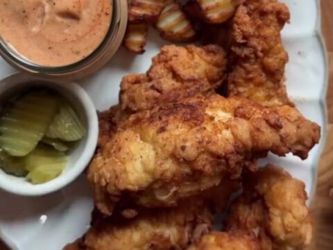 Golden-brown crispy chicken fingers piled on a plate with a bowl of pink sauce for dipping.