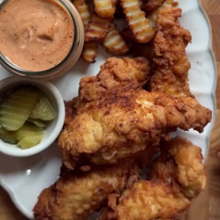 Golden-brown crispy chicken fingers piled on a plate with a bowl of pink sauce for dipping.