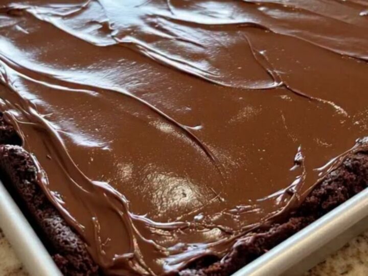 A close-up of a classic Texas sheet cake in a metal pan, topped with a smooth, glossy chocolate fudge icing.