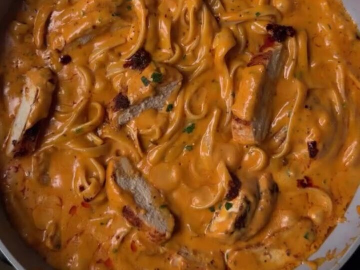 A fork twirling creamy chili crisp chicken Alfredo pasta with golden chicken slices and red chili oil ribbons.