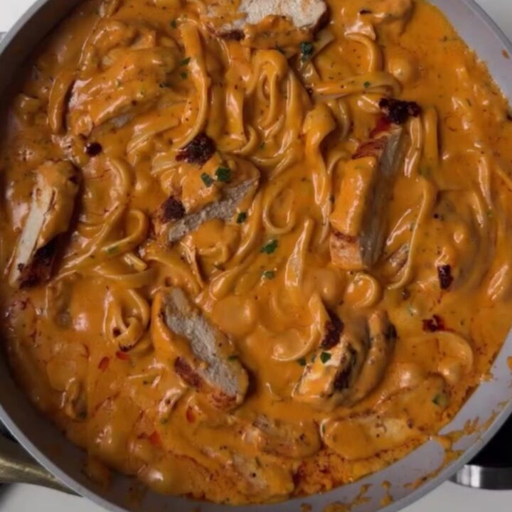 A fork twirling creamy chili crisp chicken Alfredo pasta with golden chicken slices and red chili oil ribbons.