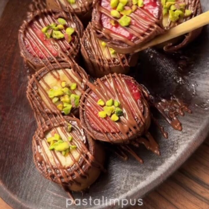 Chocolate crepe sushi rolls drizzled with white chocolate and sprinkled with pistachios on a slate platter.