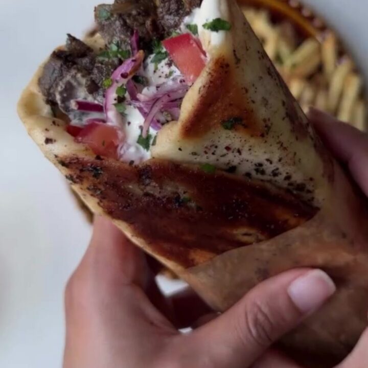 Crispy pomegranate-glazed ribeye shawarma wrap sliced in half, showing juicy beef, fries, and pink onions.