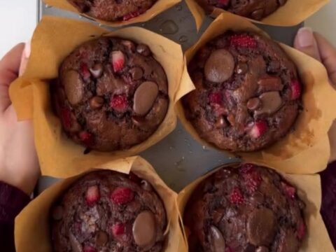 A tall chocolate muffin split open, showing molten chocolate chips and roasted strawberry pieces inside, with a crackly top.