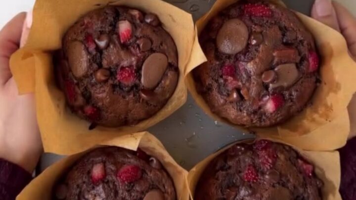 A tall chocolate muffin split open, showing molten chocolate chips and roasted strawberry pieces inside, with a crackly top.