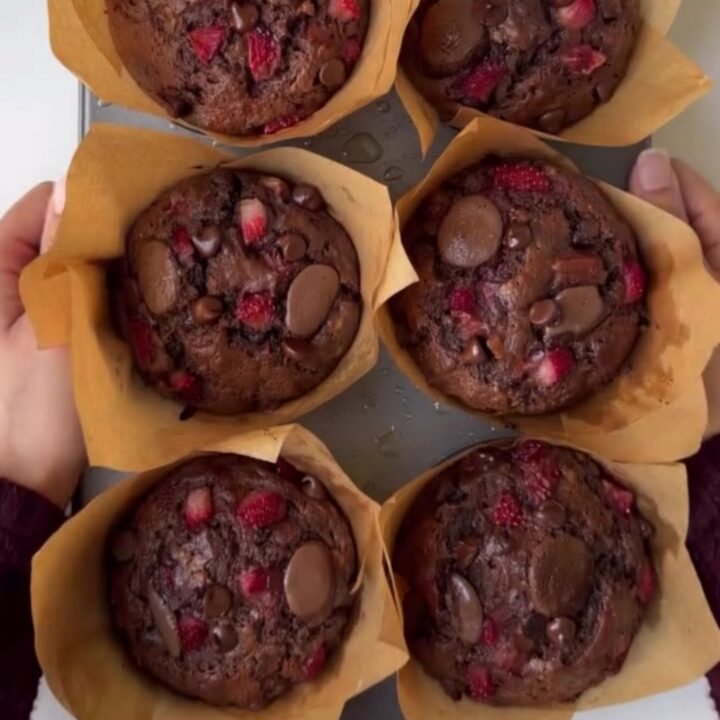 A tall chocolate muffin split open, showing molten chocolate chips and roasted strawberry pieces inside, with a crackly top.