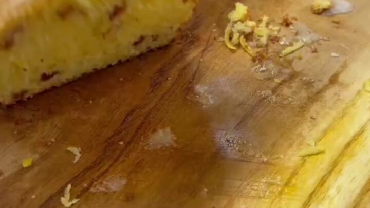 Whole lemon loaf with white glaze on a wooden board, sliced to show yellow crumb