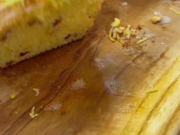 Whole lemon loaf with white glaze on a wooden board, sliced to show yellow crumb