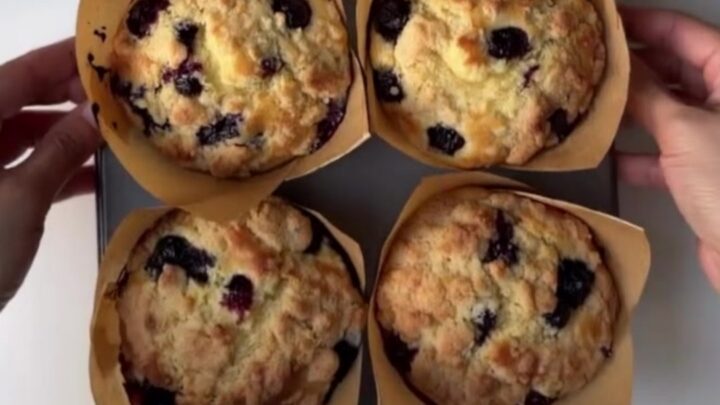 Tall blueberry lemon crumb muffins on a cooling rack, golden streusel topping, one broken open showing juicy blueberries.