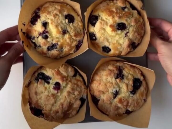Tall blueberry lemon crumb muffins on a cooling rack, golden streusel topping, one broken open showing juicy blueberries.