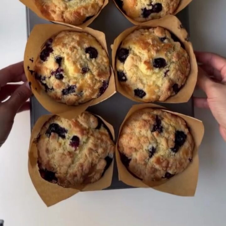 Tall blueberry lemon crumb muffins on a cooling rack, golden streusel topping, one broken open showing juicy blueberries.