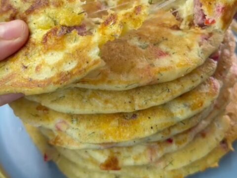 A stack of golden brown savory pancakes with red peppers and dill, steam rising as one is broken open to reveal melted feta.