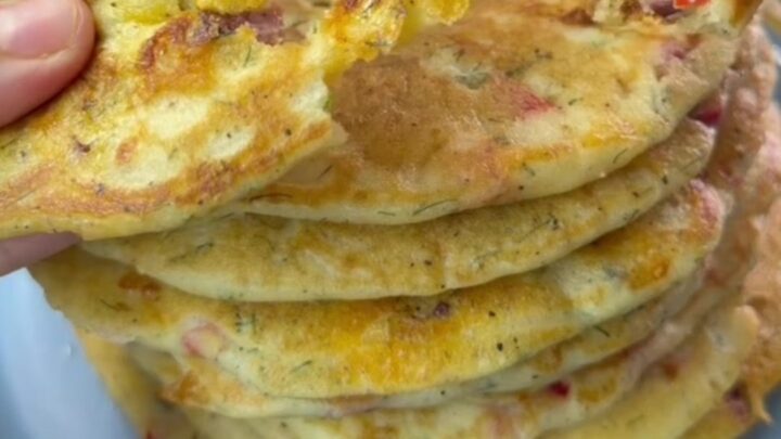 A stack of golden brown savory pancakes with red peppers and dill, steam rising as one is broken open to reveal melted feta.
