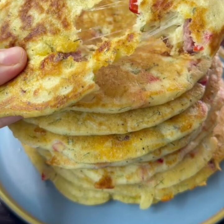 A stack of golden brown savory pancakes with red peppers and dill, steam rising as one is broken open to reveal melted feta.