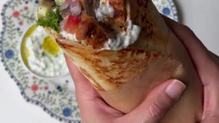 Sliced Greek chicken wrap showing layers of charred chicken, romaine, tomatoes, and tzatziki in a warm tortilla.