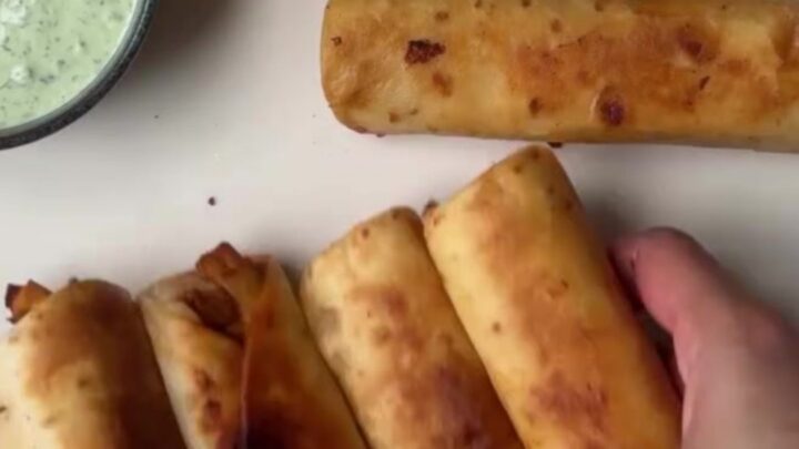 Crispy golden chicken taquitos arranged on a platter with a bowl of vibrant green crema for dipping