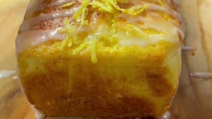 Whole lemon blender loaf cake with glossy white glaze and fresh lemon zest on top, sliced to show tender yellow crumb.