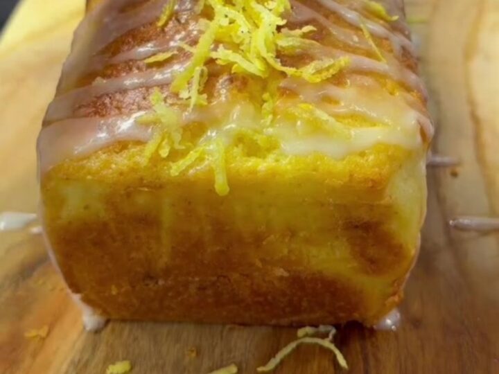 Whole lemon blender loaf cake with glossy white glaze and fresh lemon zest on top, sliced to show tender yellow crumb.