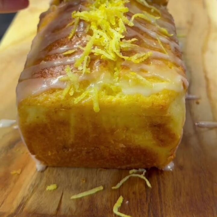 Whole lemon blender loaf cake with glossy white glaze and fresh lemon zest on top, sliced to show tender yellow crumb.