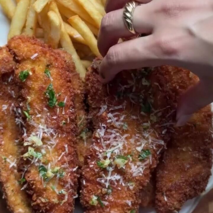 Crispy garlic Parmesan chicken tenders piled on a plate with a bowl of chili sauce for dipping.