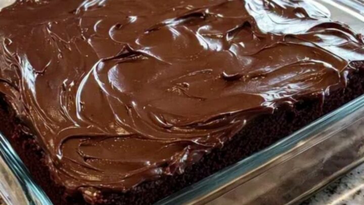 A close-up of a moist chocolate cake with glossy fudge frosting in a square glass dish.