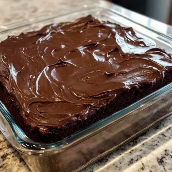 A close-up of a moist chocolate cake with glossy fudge frosting in a square glass dish.