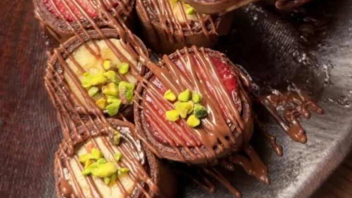Chocolate crepe sushi rounds with colorful fruit centers on a plate with a bowl of chocolate syrup and wooden chopsticks.