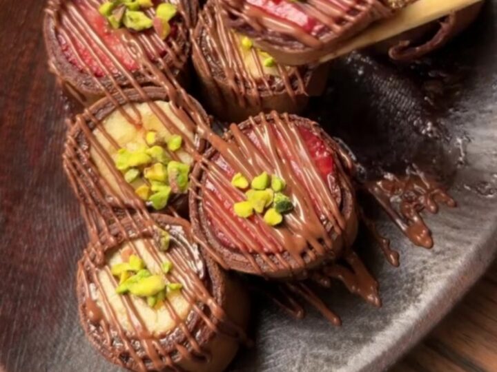 Chocolate crepe sushi rounds with colorful fruit centers on a plate with a bowl of chocolate syrup and wooden chopsticks.