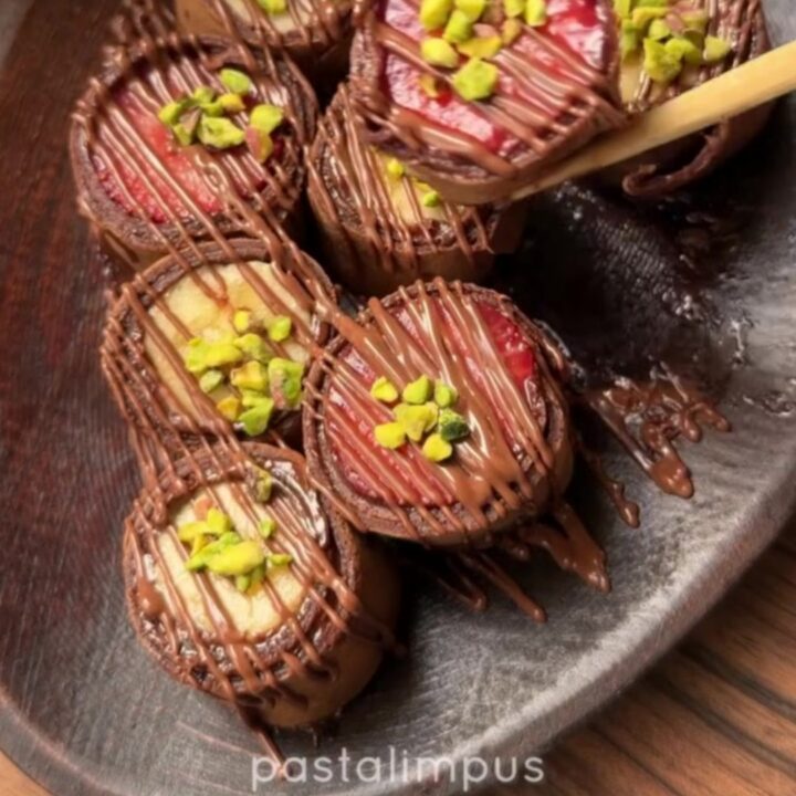 Chocolate crepe sushi rounds with colorful fruit centers on a plate with a bowl of chocolate syrup and wooden chopsticks.