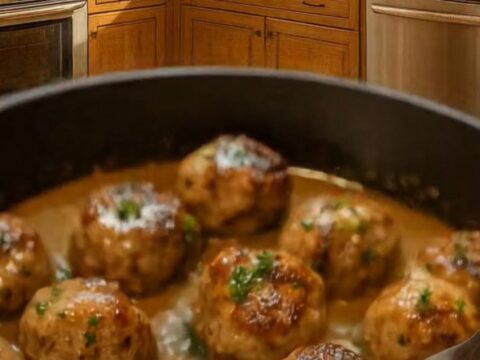 A close-up of tender, golden-brown meatballs in a dark skillet, covered in a rich, velvety cream sauce and fresh parsley.