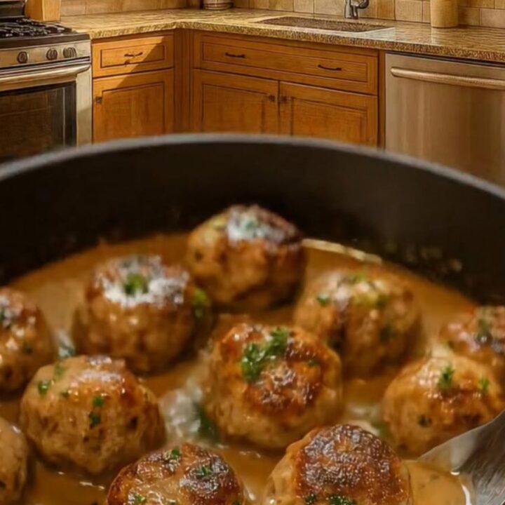 A close-up of tender, golden-brown meatballs in a dark skillet, covered in a rich, velvety cream sauce and fresh parsley.