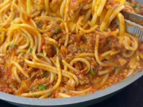 A forkful of spaghetti with melted mozzarella cheese pull, coated in tomato meat sauce, garnished with basil.