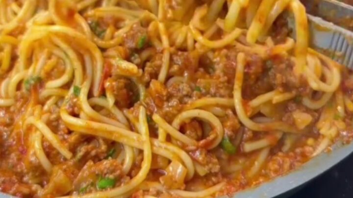 A forkful of spaghetti with melted mozzarella cheese pull, coated in tomato meat sauce, garnished with basil.