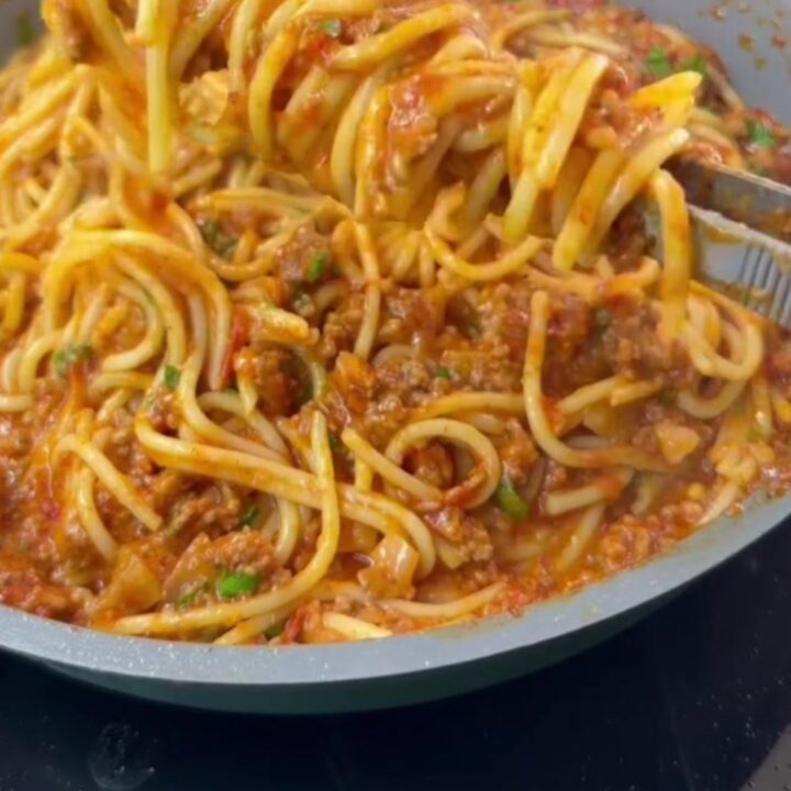 A forkful of spaghetti with melted mozzarella cheese pull, coated in tomato meat sauce, garnished with basil.