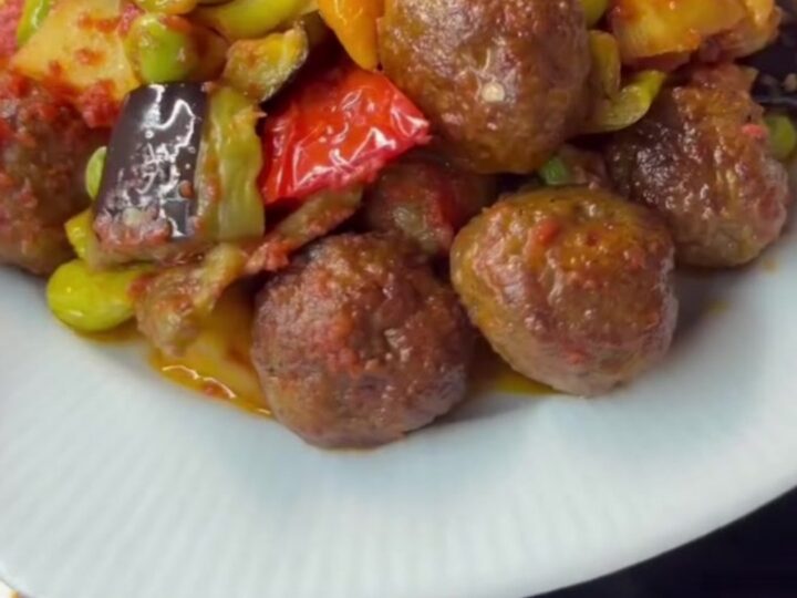 Baked meatballs with fried eggplant and potatoes in a glass dish, topped with peas and peppers, steam rising.