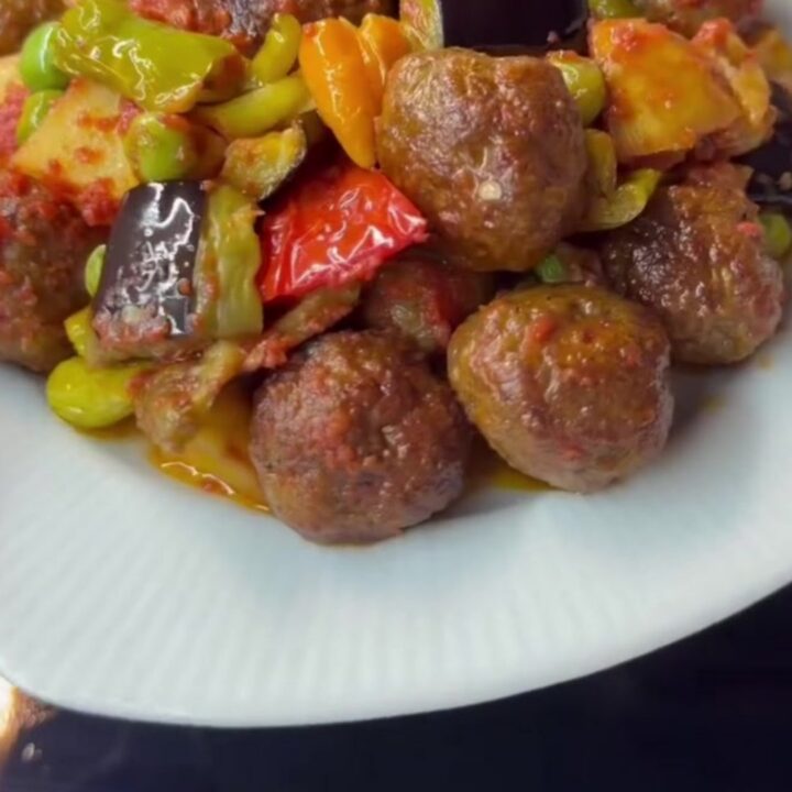 Baked meatballs with fried eggplant and potatoes in a glass dish, topped with peas and peppers, steam rising.